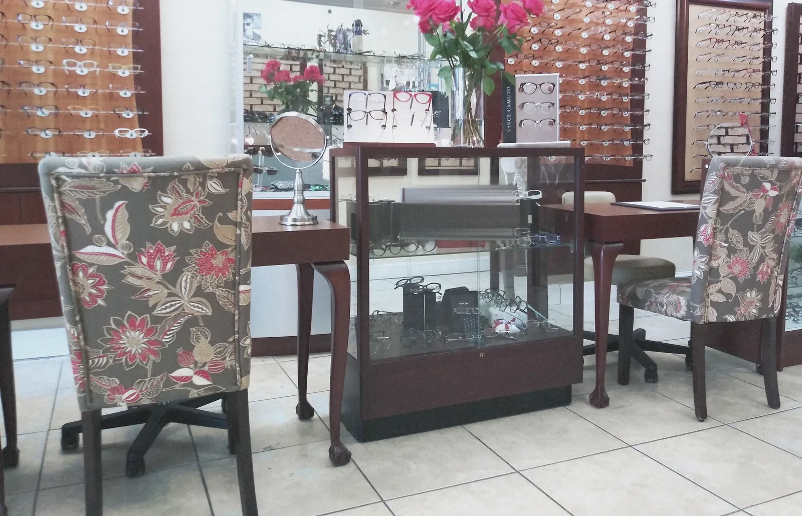 La sala de la óptica: dos sillas tapizadas con flores, una vitrina de cristal con monturas y rosas rojas al fondo
